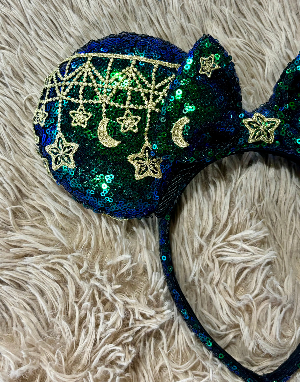 A close-up of a pair of sequined ears with a dark blue and green iridescent base, decorated with gold sequined stars, crescent moons, and a web pattern, resting on a fluffy cream faux fur surface.