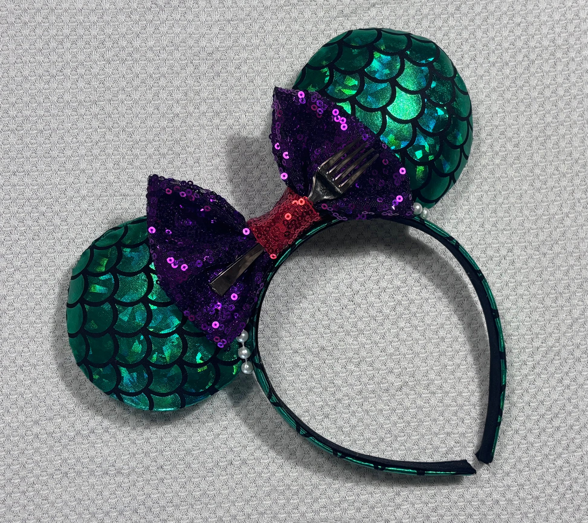 Mermaid-themed headband with teal scales and a purple sequin bow with a red middle and a dinglehopper middle piece. Pearl garland trim along inner ears m on a matching mermaid scales headband