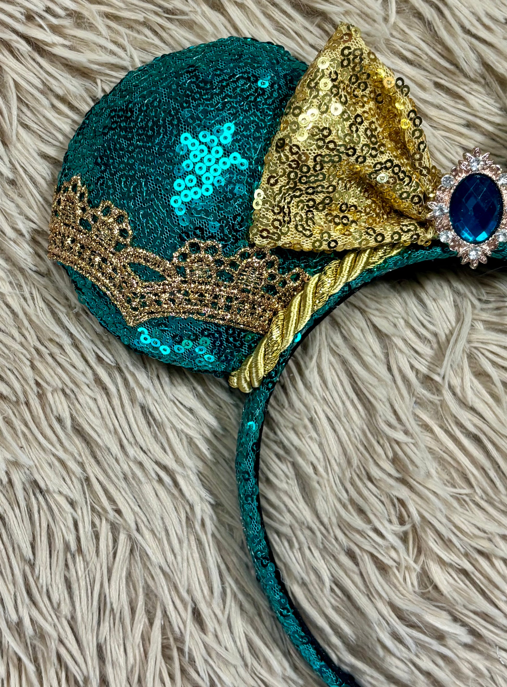 A close-up of a sequined headband featuring a teal green sequin-covered cup, a large gold sequin bow, a gold lace trim, a twisted gold ribbon detail, and a blue gemstone setting on a beige shag rug.