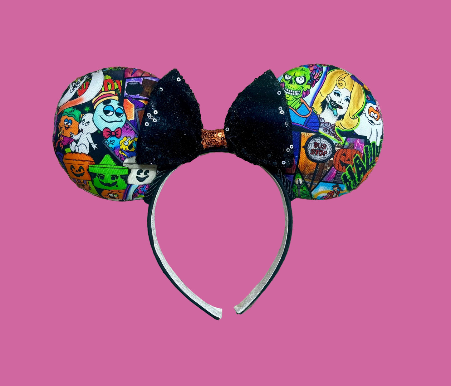 An ear headband accessory featuring a colorful Halloween-themed print on the outer ears and a black glitter bow in the center with black twisted trim along the inner ears.