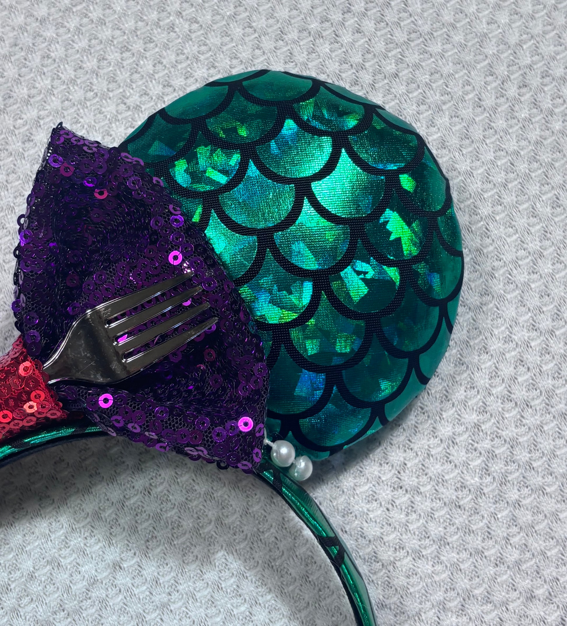 Close up of other front ear. Mermaid-themed headband with teal scales and a purple sequin bow with a red middle and a dinglehopper middle piece. Pearl garland trim along inner ears m on a matching mermaid scales headband