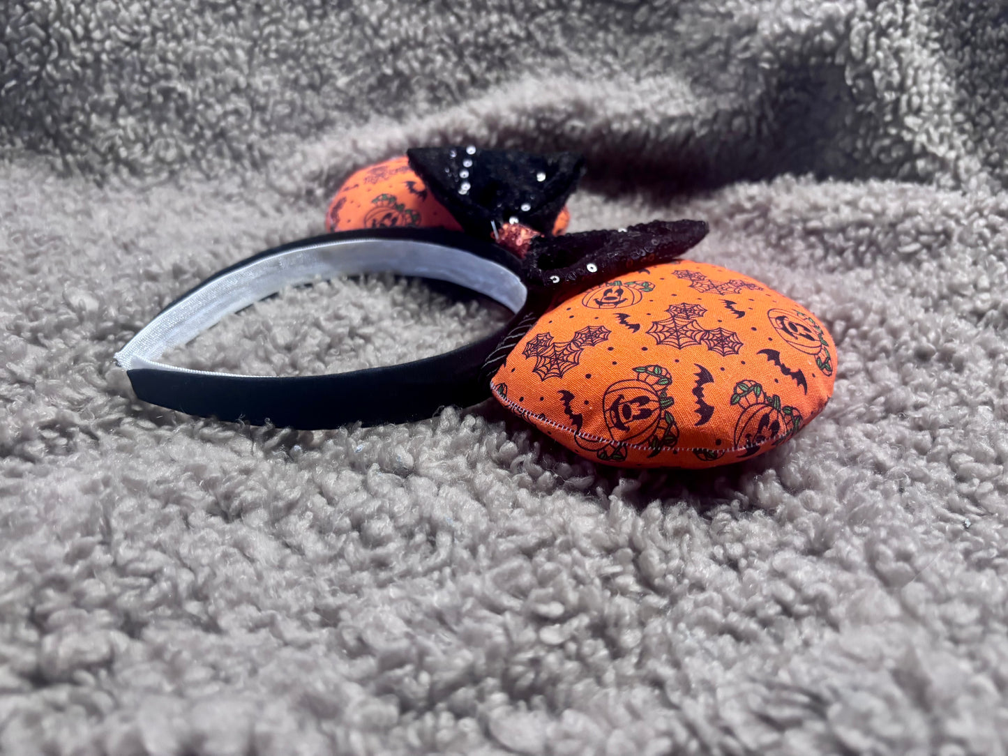 Halloween mouse ears with the fabric on both the front and back. It's orange with bats, mouse shaped pumpkins and spider webs. The headband is black universal sized and fits most. The set has a black with orange middle sequin bow. Item is shown on a brown furry background.