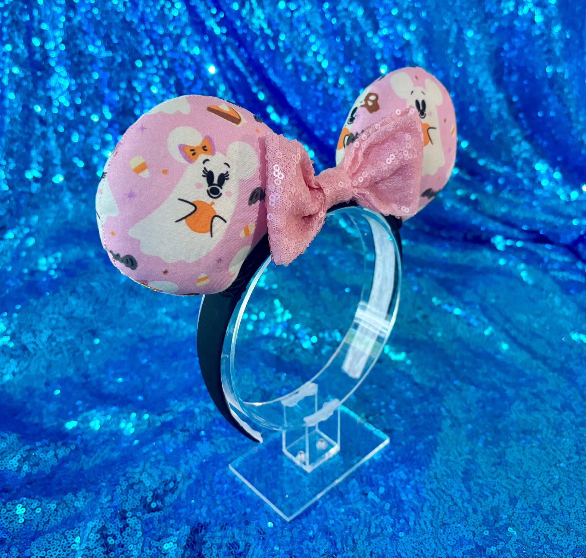 Side angled view of the headband on a clear stand. Handmade and ready to ship Halloween Pink Ghosts Mouse ears with the fabric with mouse shaped ghosts on pink fabric is on the ears, front and back and a light pink sequin bow. The headband is a universal sized headband and has black poly cotton fabric on it.