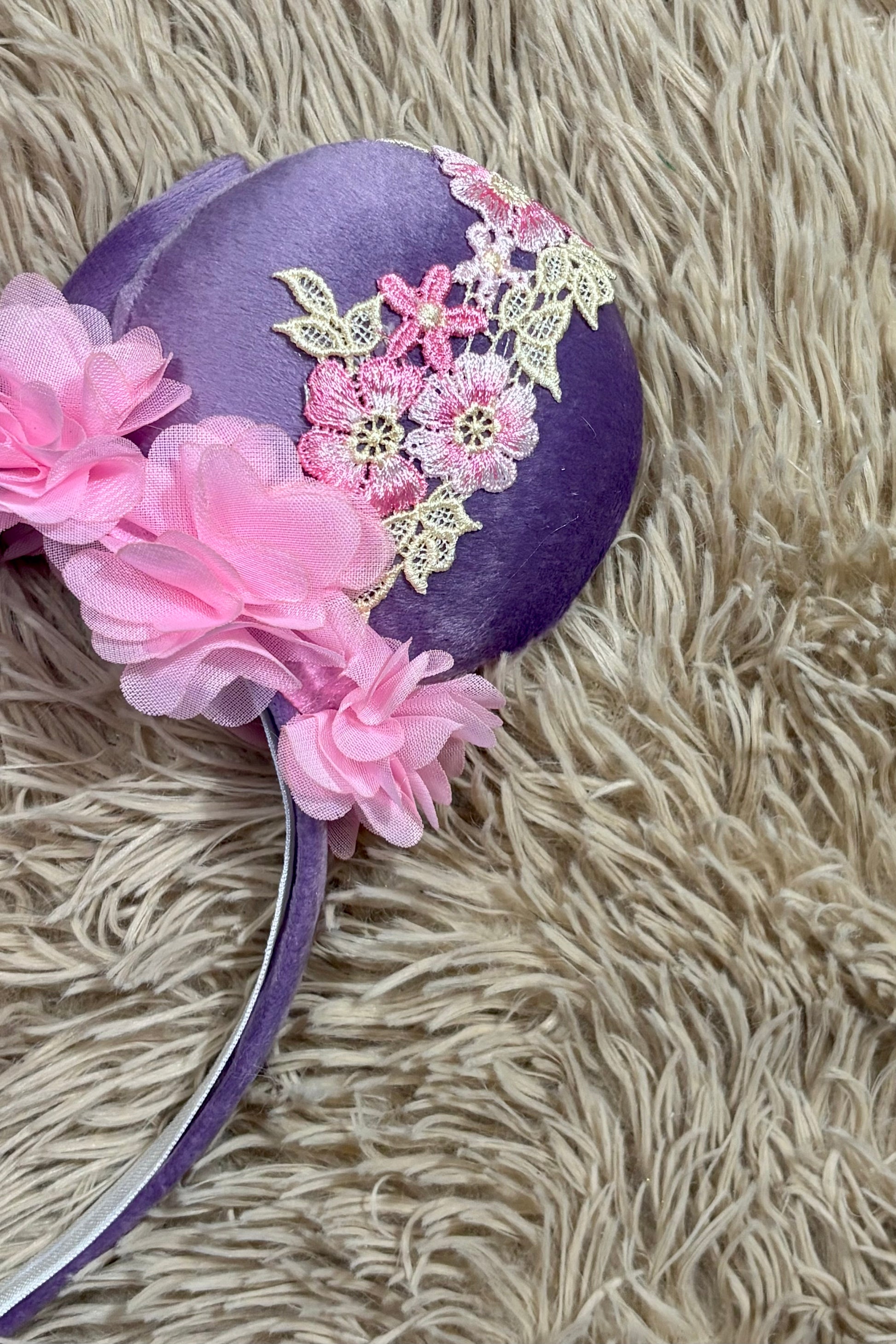 A purple fabric headband featuring pink fabric flowers and a pink floral lace applique, displayed on a beige shag rug.