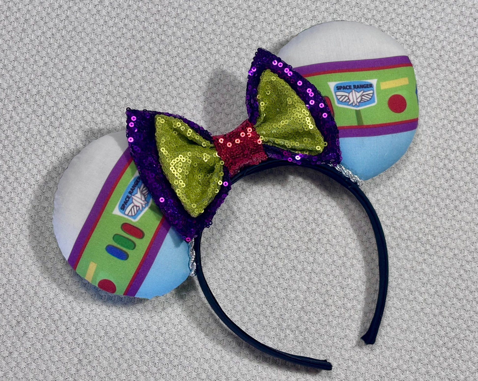 Space Ranger Mouse Ear headband with new improved bow. Handmade and ready to ship with Space ranger fabric on both the front and the back of mouse ears. The set has a silver twisted trim along the inner ears and a new sequin bow that is purple, lime green and red in sequin. Ears are on a black poly cotton universal sized headband.