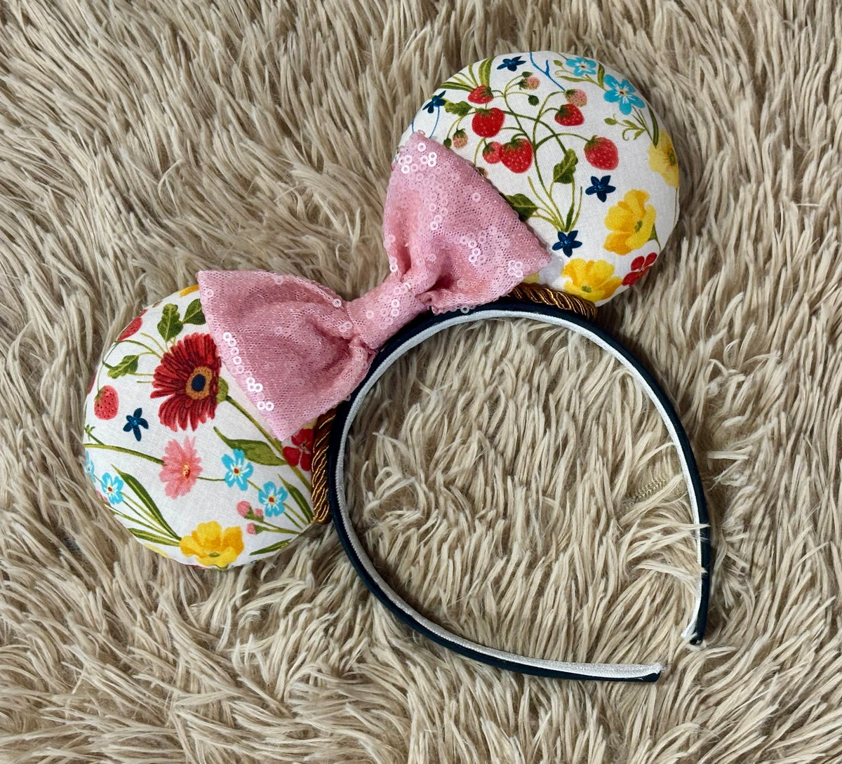A handmade ear headband featuring a white background with colorful floral and strawberry patterns, a large pink sequined bow, and a black headband, laid flat on a beige shag rug.