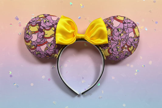 Figment cartoon inspired fabric is on the Mouse ears. On a black headband with a yellow satin bow. Shown on a colorful background