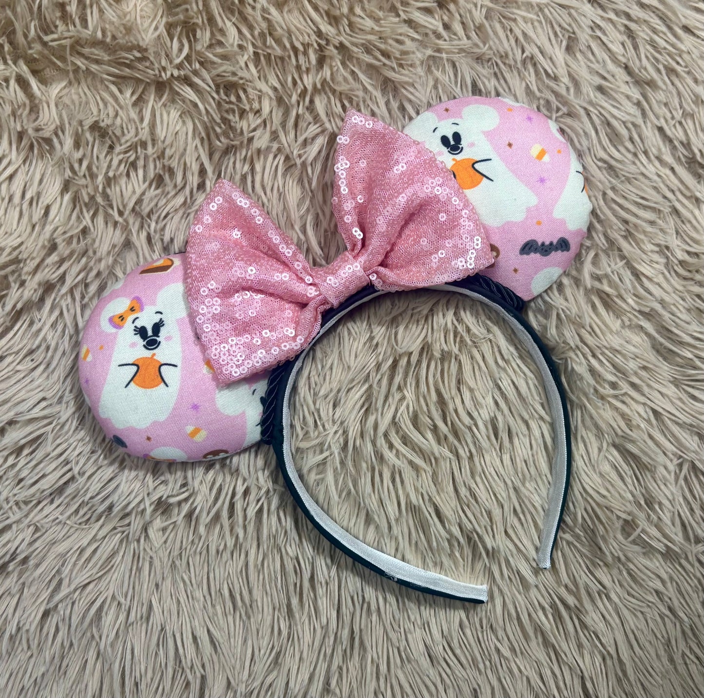 An ear headband featuring a pink sequined bow in the center and pink ghost-patterned ears.