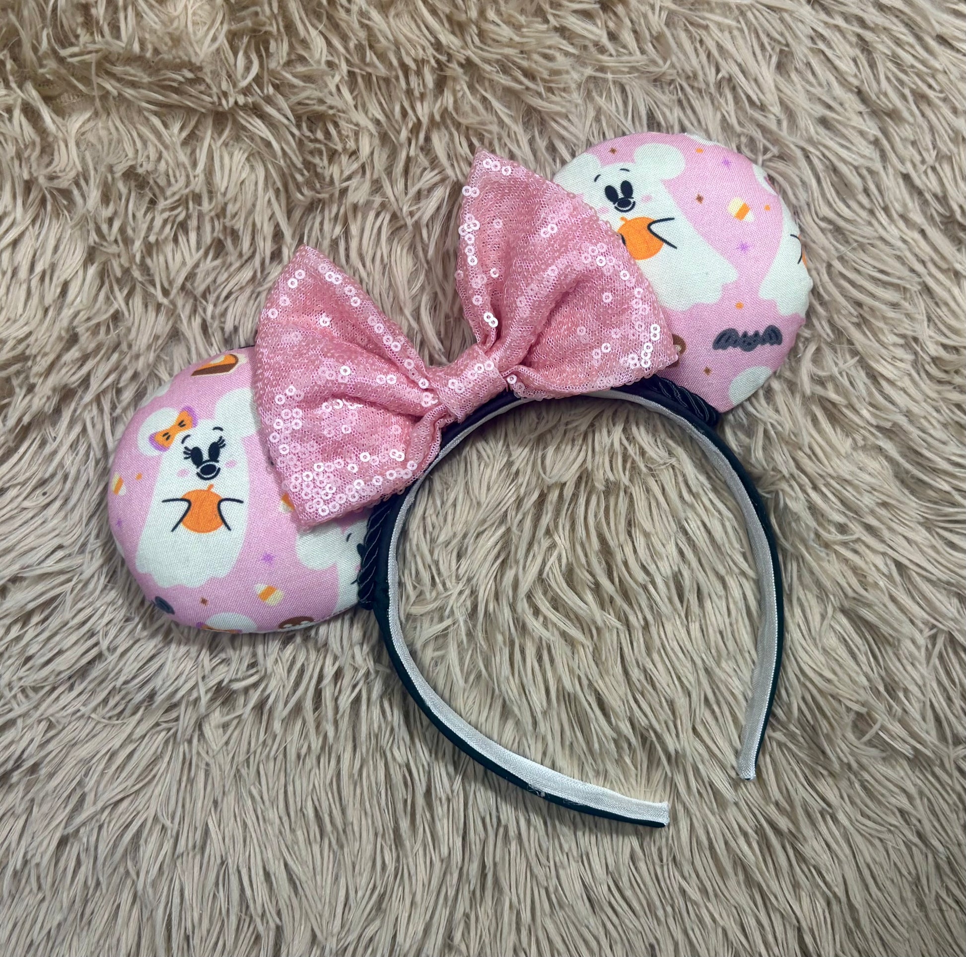 An ear headband featuring a pink sequined bow in the center and pink ghost-patterned ears.