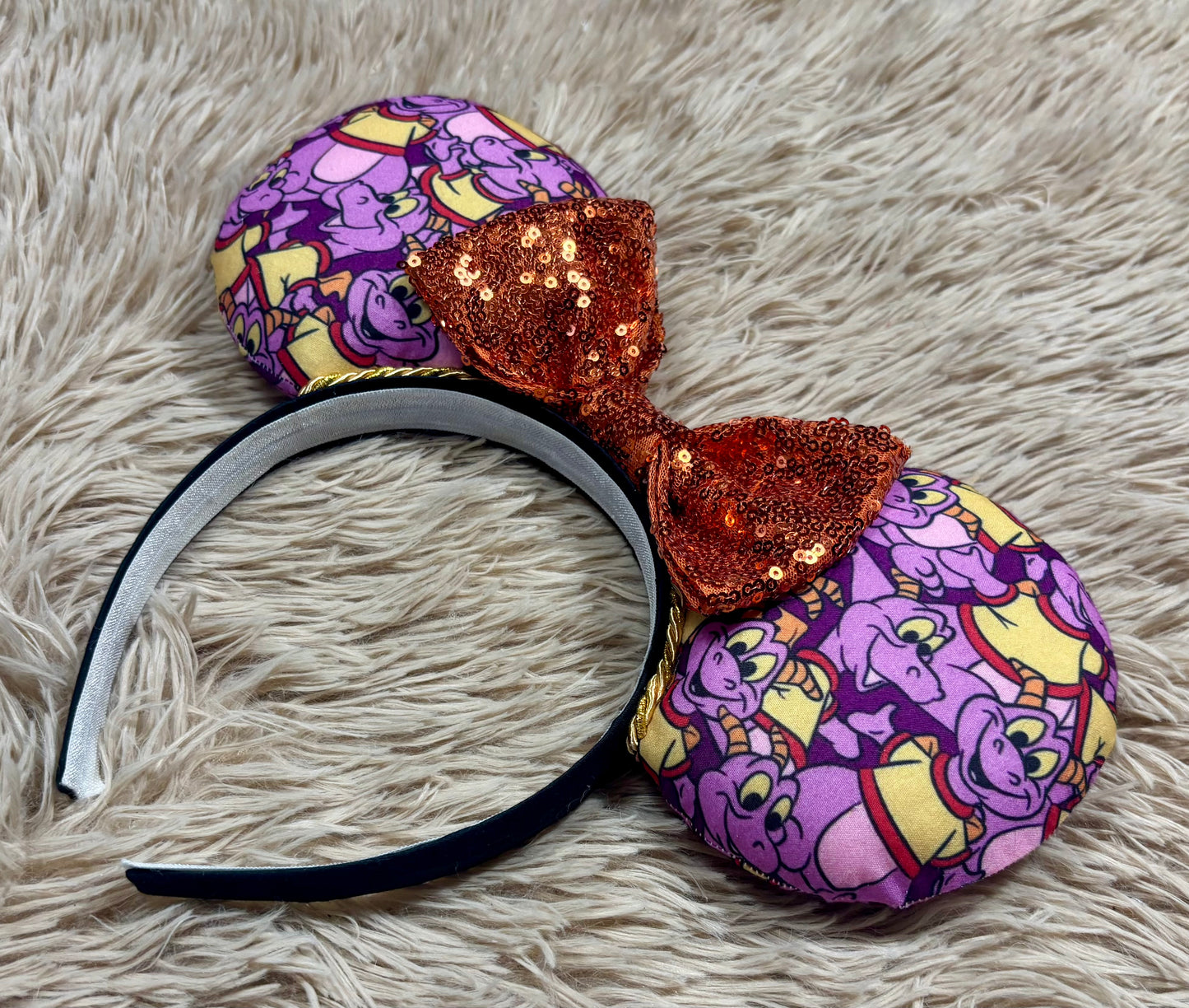 A pair of dragon style ears with a black headband, featuring a purple and yellow cartoon pattern on the ears and an orange sequined bow in the center