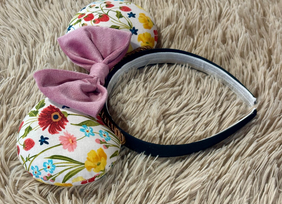 A headband with two round floral ears and a large dusty pink bow, displayed on a beige shag rug.