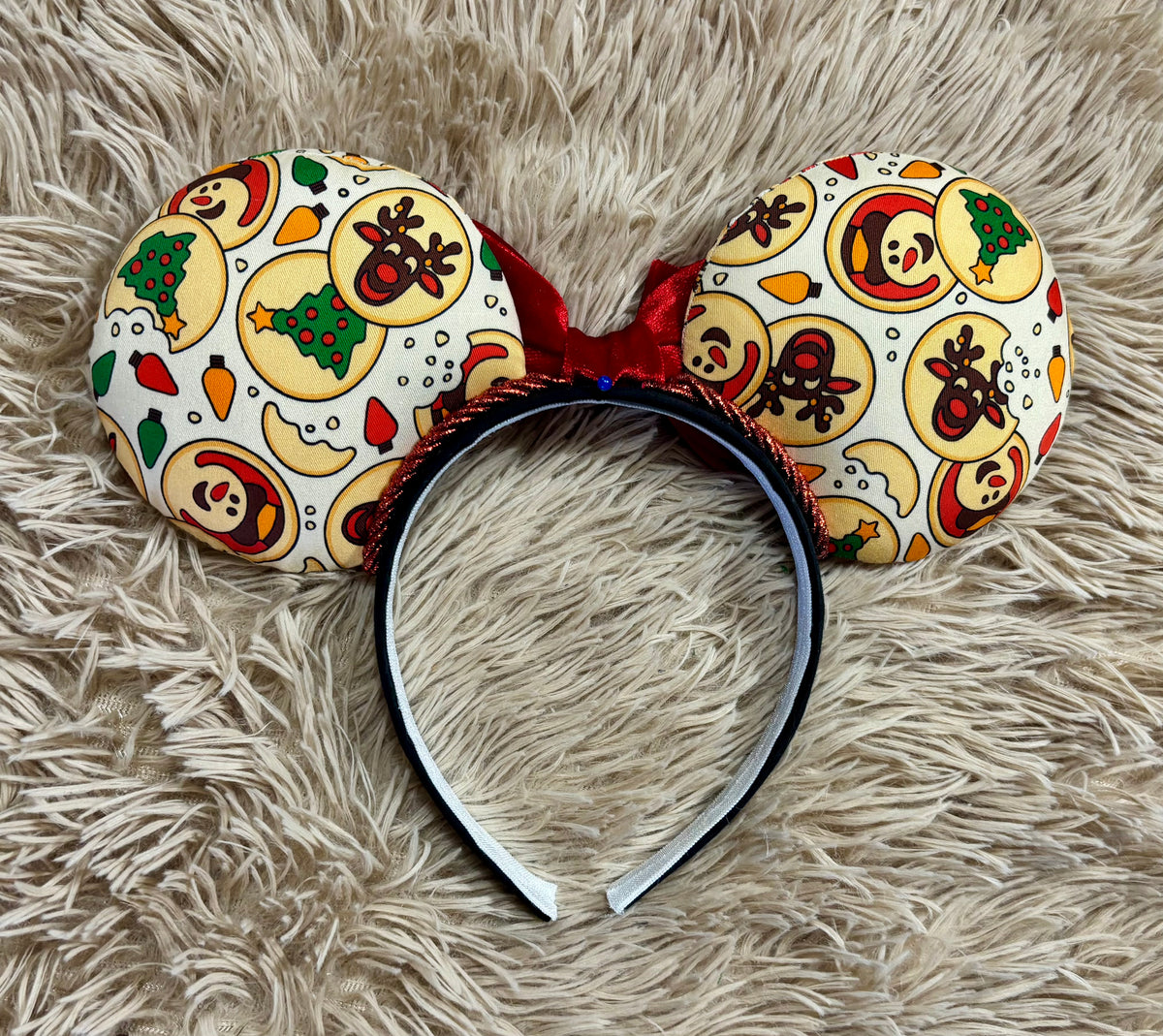 A pair of headband ears with a Christmas-themed pattern of gingerbread cookies, reindeer, and holiday motifs, resting on a beige faux fur surface.