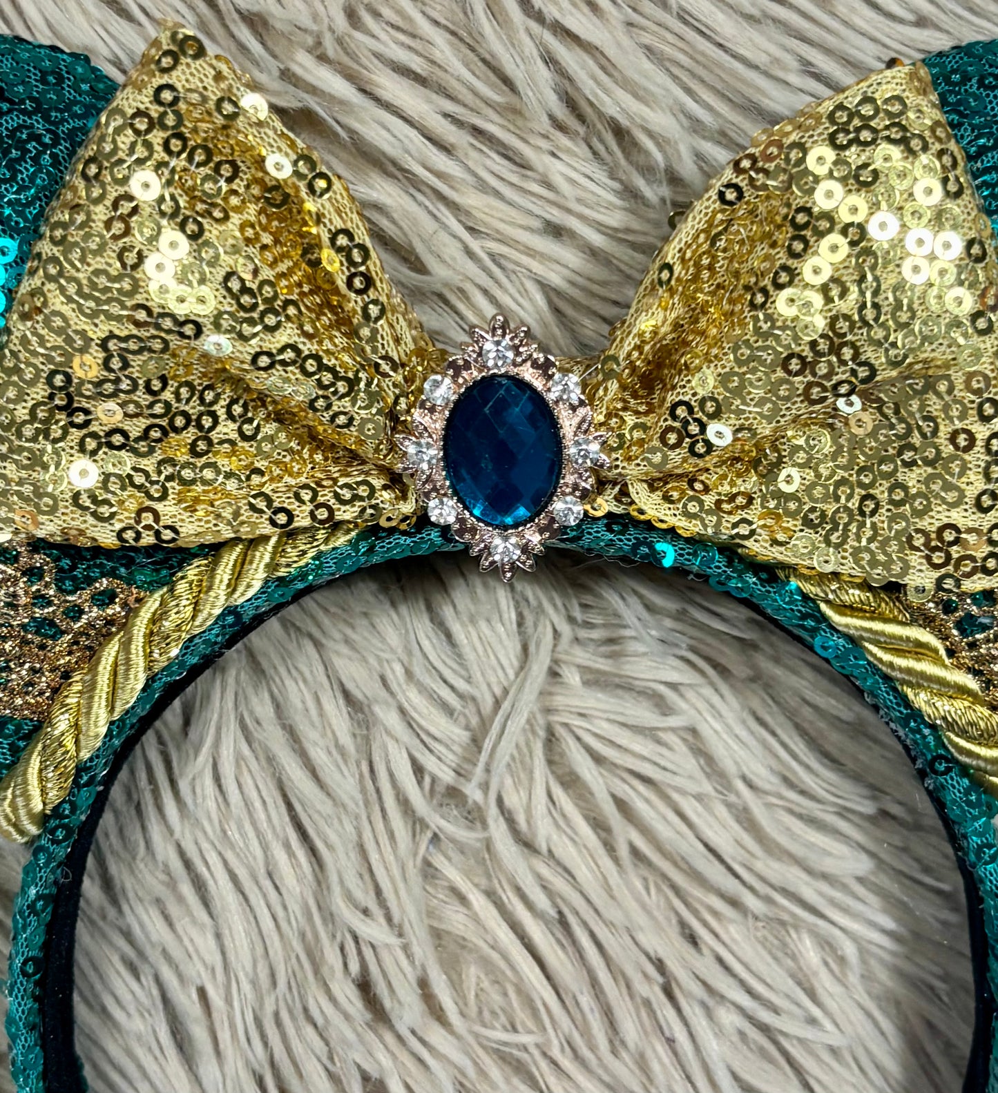 A close-up of a decorative headband featuring a large bow made of gold sequins and teal trim, with a central blue oval gemstone surrounded by small clear rhinestones, set against a shaggy light gray background.