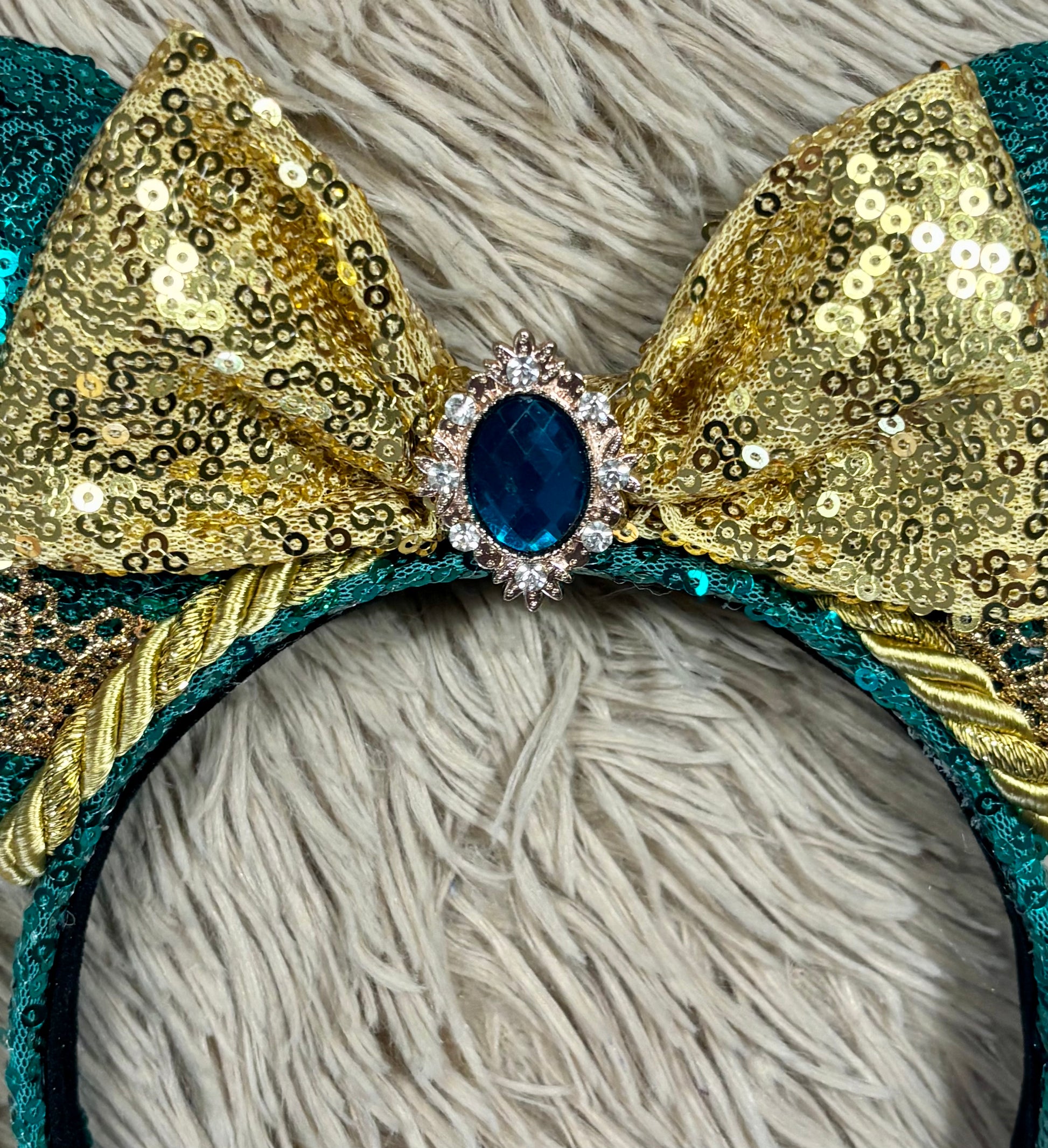 A close-up of a decorative headband featuring a large bow made of gold sequins and teal trim, with a central blue oval gemstone surrounded by small clear rhinestones, set against a shaggy light gray background.