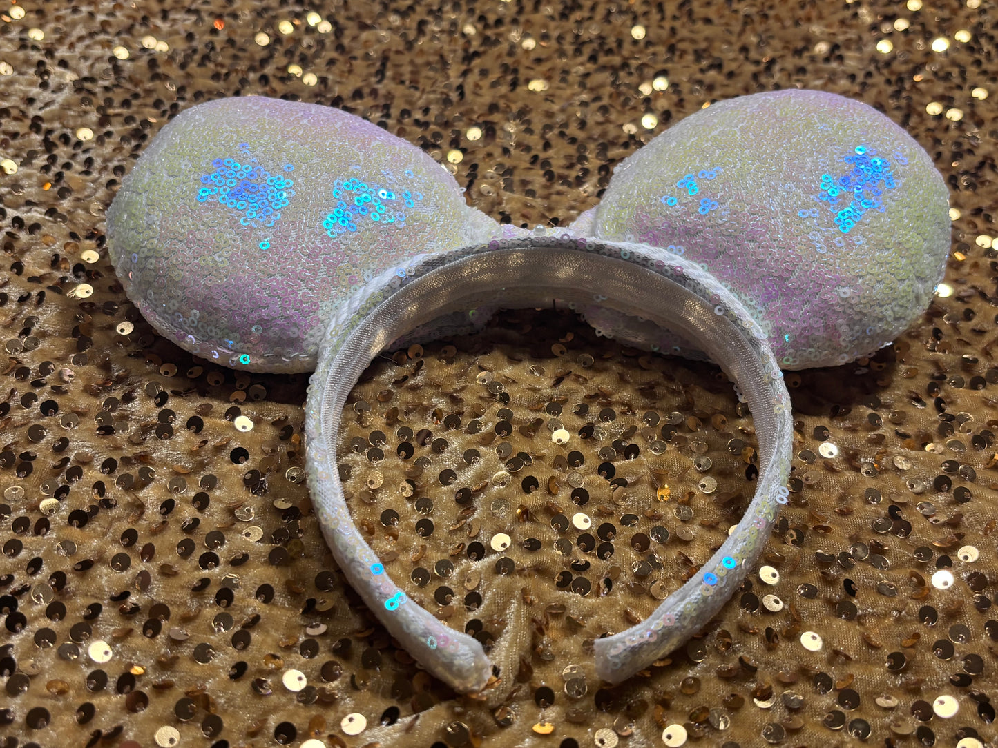 White Iridescent mouse ears with the fabric on both the front and back. The fabric is on the headband as well and is universal sized and fits most. The set has a matching iridescent bow sequin bow. Item is shown on a gold sequin background.