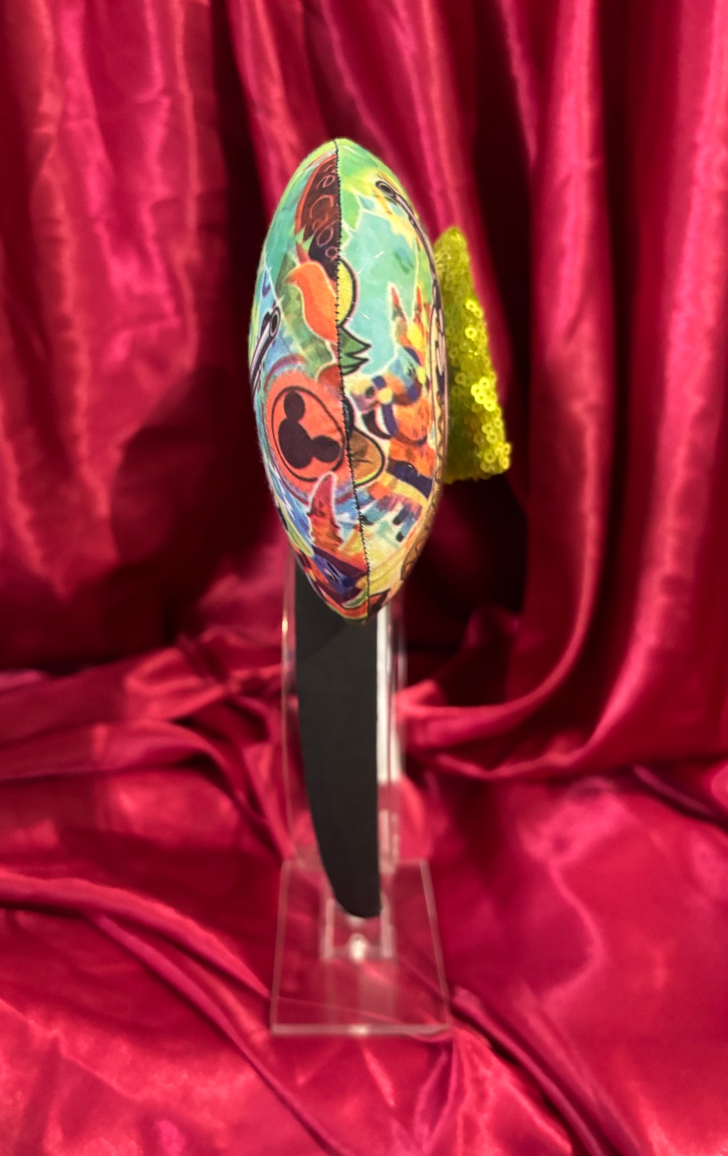 Side view of Colorful Handmade mouse ears with a green bow on a red satin background with duck cartoon images on the ears