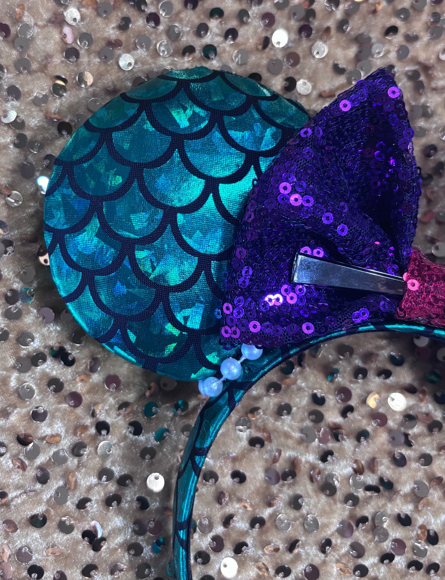 Mermaid-themed headband with teal scales and a purple sequin bow with a red middle and a dinglehopper middle piece. Pearl garland trim along inner ears m on a matching mermaid scales headband.