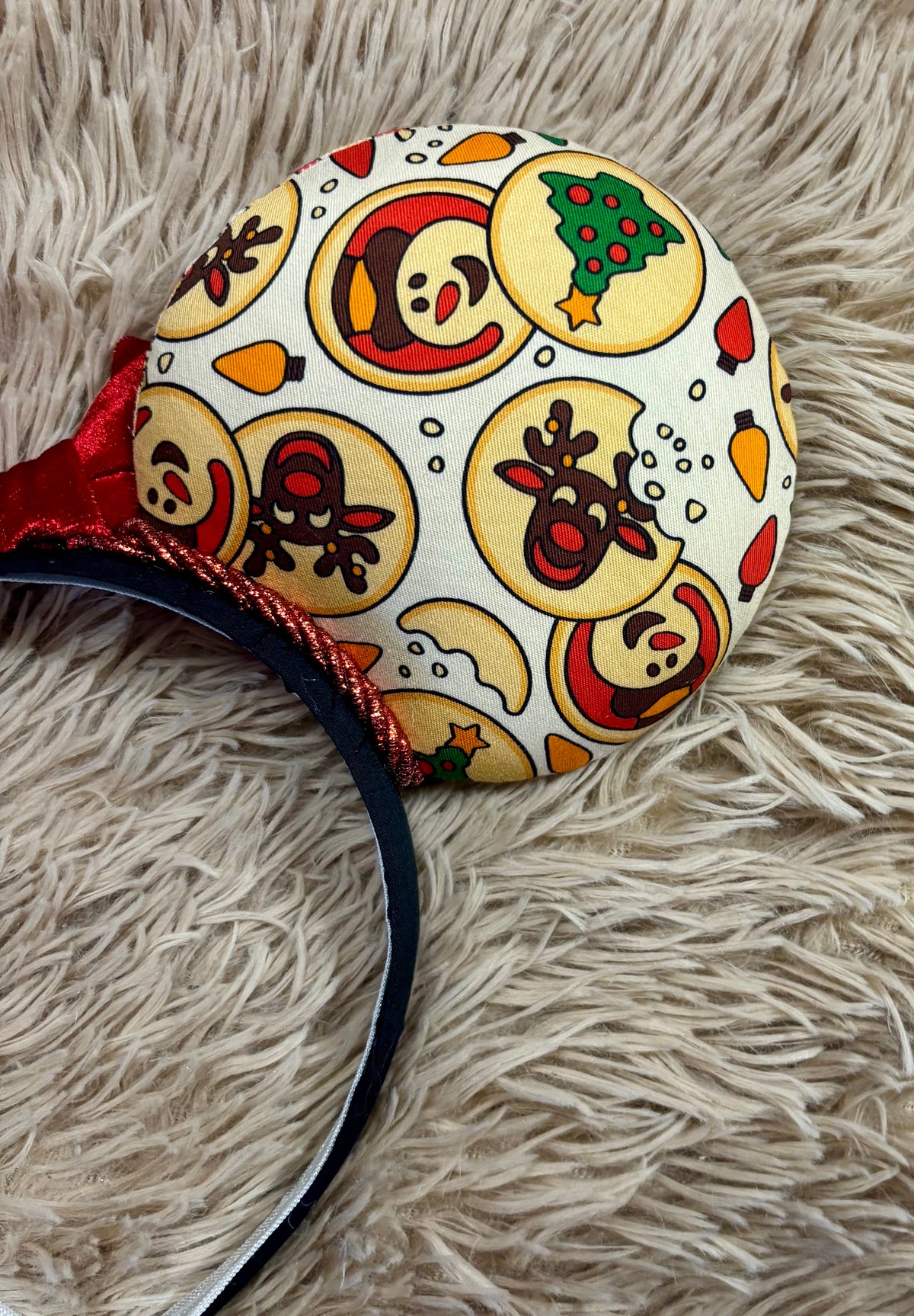 A close-up of a round headband featuring a festive Christmas cookie print with a penguin, reindeer, snowman, and Christmas tree on a cream background, resting on a beige faux fur surface.