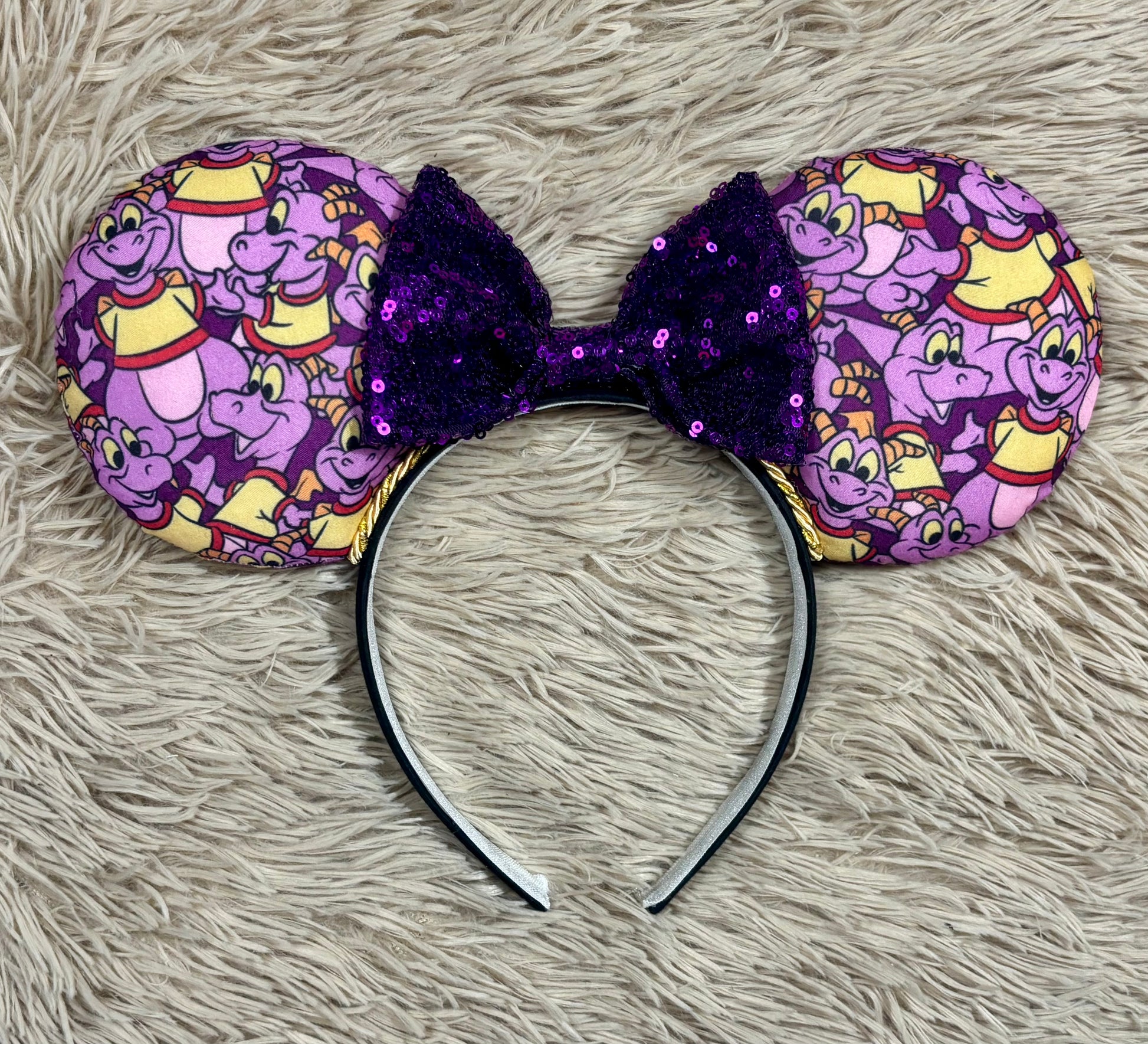 A pair of dragon style ears with a black headband, featuring a purple and yellow cartoon pattern on the ears and a dark purple sequined bow in the center