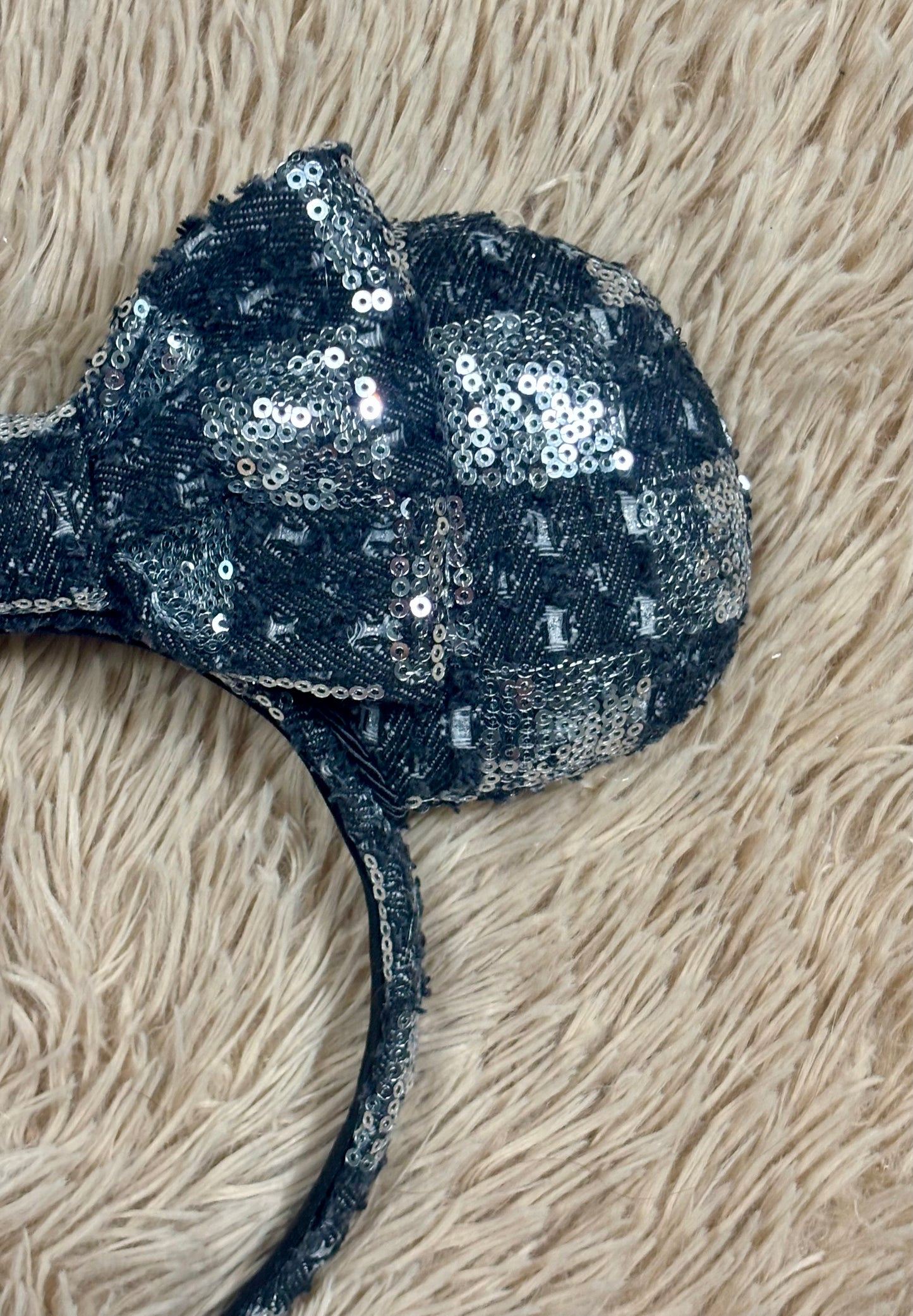 A close-up of a black and silver checkered sequined headband with a large bow, laid flat on a beige shag rug.