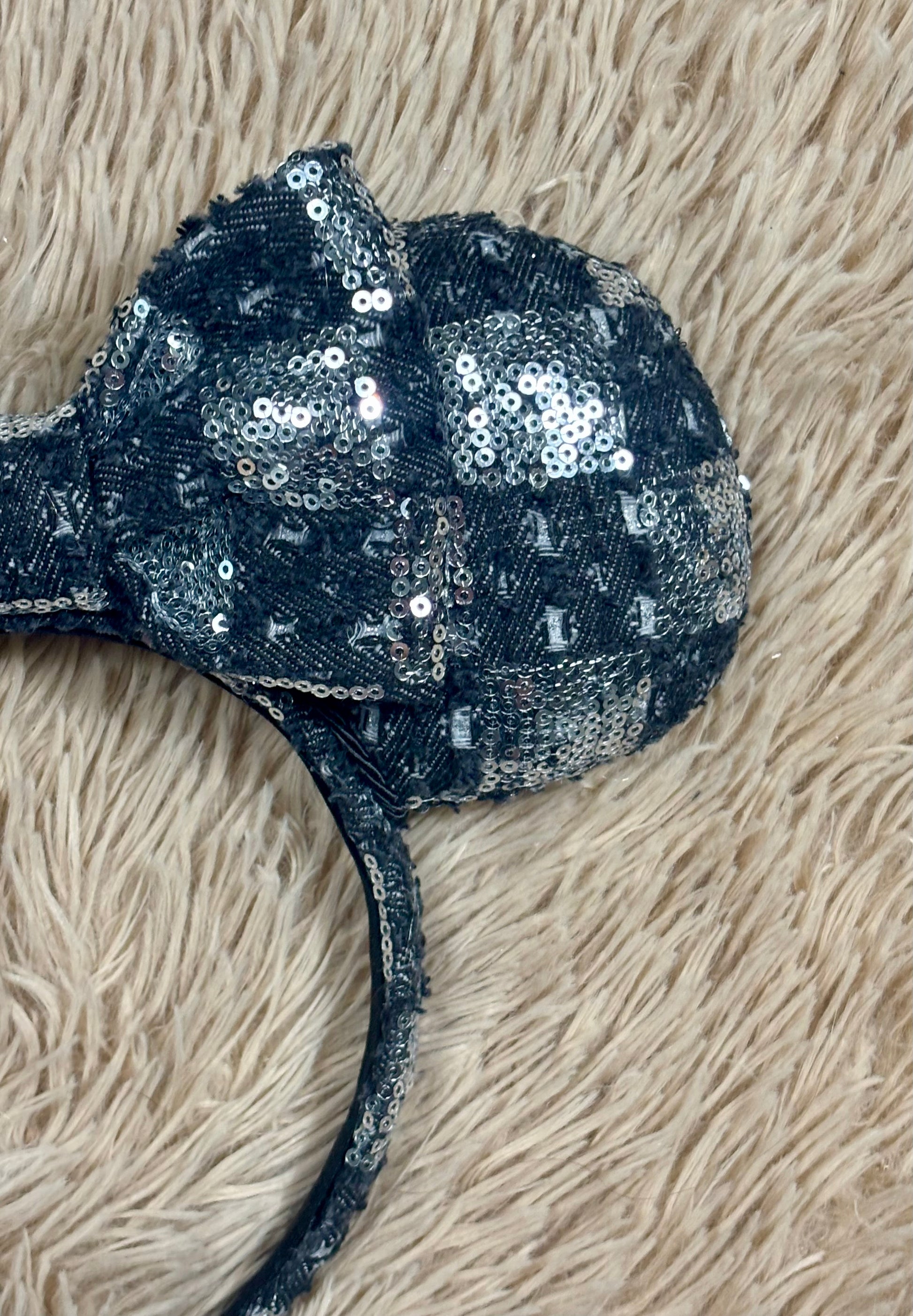 A close-up of a black and silver checkered sequined headband with a large bow, laid flat on a beige shag rug.