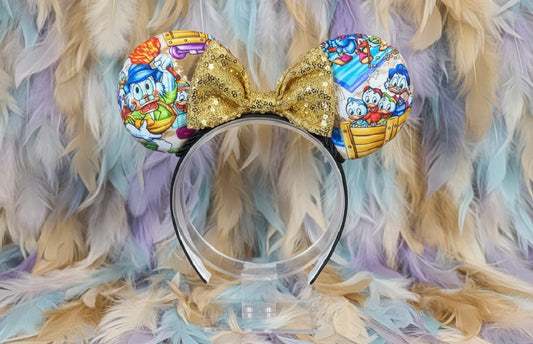 Duck Tales themed mouse ears with a gold sequin bow on a black headband. Shown on a feathered background