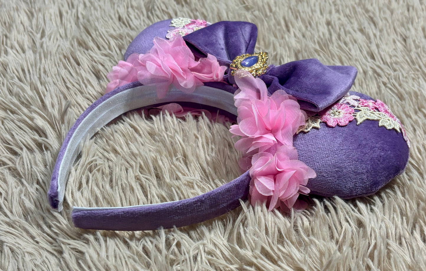 A purple velvet headband featuring a large bow, pink fabric flowers, lace trim, and a decorative jewel, displayed on a beige shag rug.
