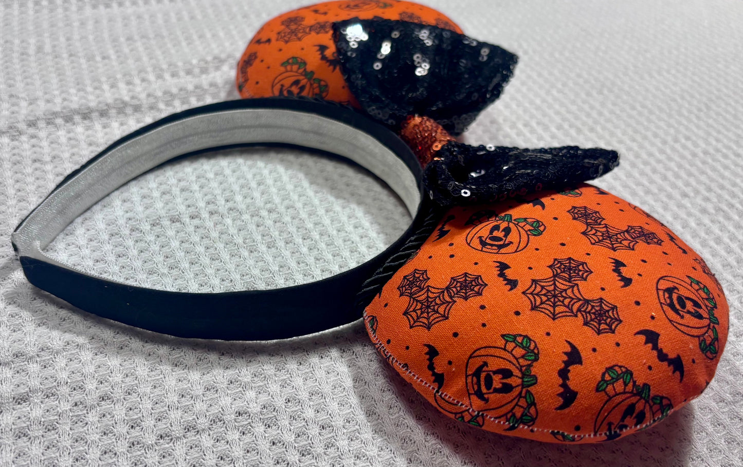Halloween mouse ears with the fabric on both the front and back. It's orange with bats, mouse shaped pumpkins and spider webs. The headband is black universal sized and fits most. The set has a black with orange middle sequin bow. Item is shown on a white background.