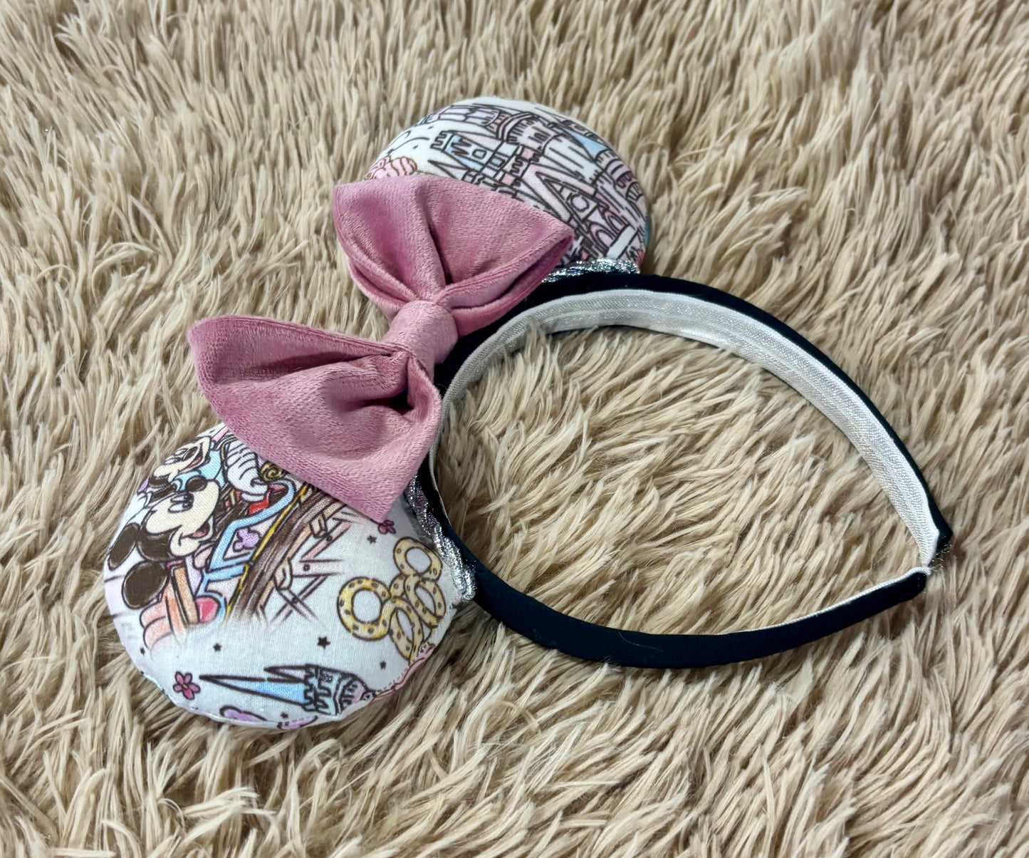 A pair of handmade ear headbands with a large dusty pink velvet bow, featuring printed park themed patterns on the ears and resting on a beige faux fur surface.