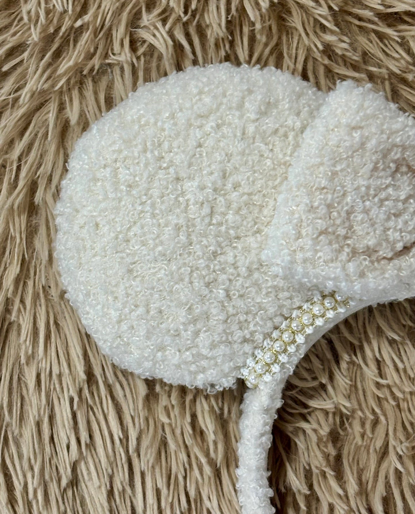 A close-up of a cream-colored, fuzzy textured headband with a small decorative band of white pearls, laid flat on a beige shaggy rug.