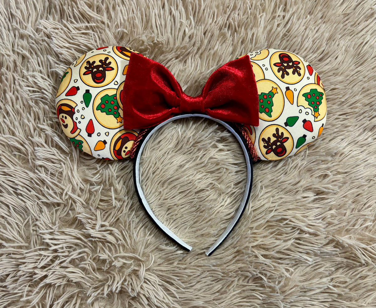 An ear headband featuring a festive Christmas print with red velvet bows, displayed on a beige faux fur background.