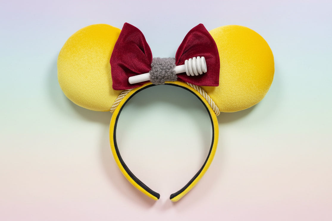 Yellow soft plush mouse ears on the ears and the headband with a red plush bow and honeycomb in middle of the bow. Shown on a light gradient background