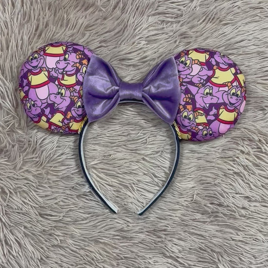 A pair of dragon style ears with a black headband, featuring a purple and yellow cartoon pattern on the ears and a dark purple sequined bow in the center