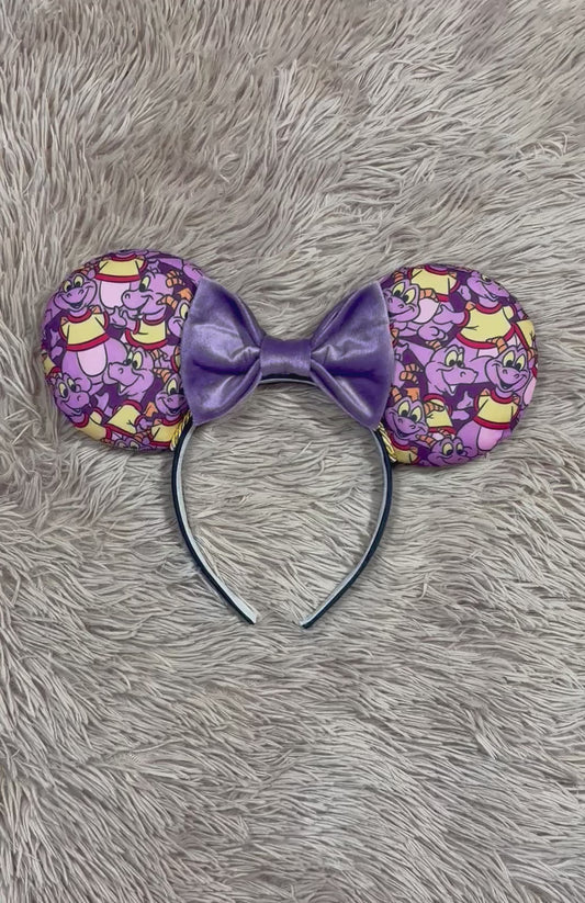 A pair of dragon style ears with a black headband, featuring a purple and yellow cartoon pattern on the ears and a dark purple sequined bow in the center