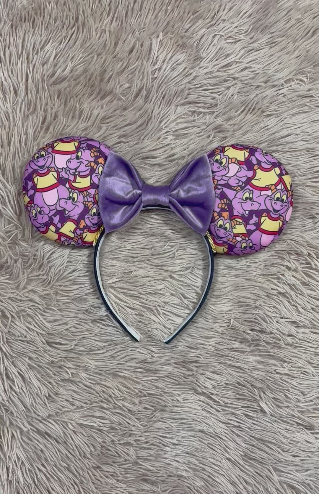 A pair of dragon style ears with a black headband, featuring a purple and yellow cartoon pattern on the ears and a dark purple sequined bow in the center