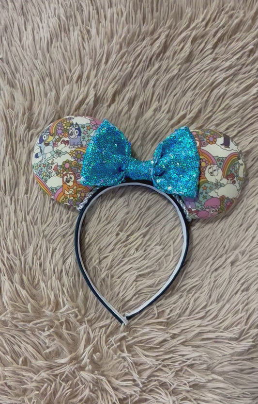 An ear headband with a colorful cartoon print and a large glittery blue bow, displayed on a beige faux-fur surface.