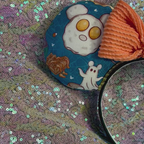 Handmade and ready to ship Fall Mouse ears with pumpkins, eggs, waffles and more shaped like mouse ears is on the front and back of mouse ears with a orange waffle print bow on a rainbow  glitter background.