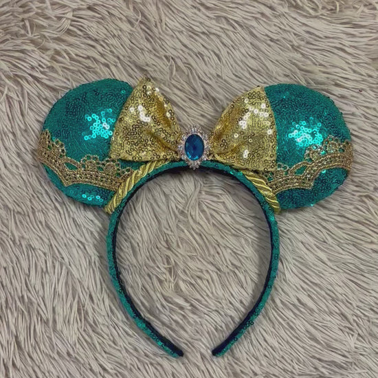A pair of sequined mouse ears headband featuring teal and gold sequins, a large gold sequin bow with a blue gemstone center, and gold lace trim.