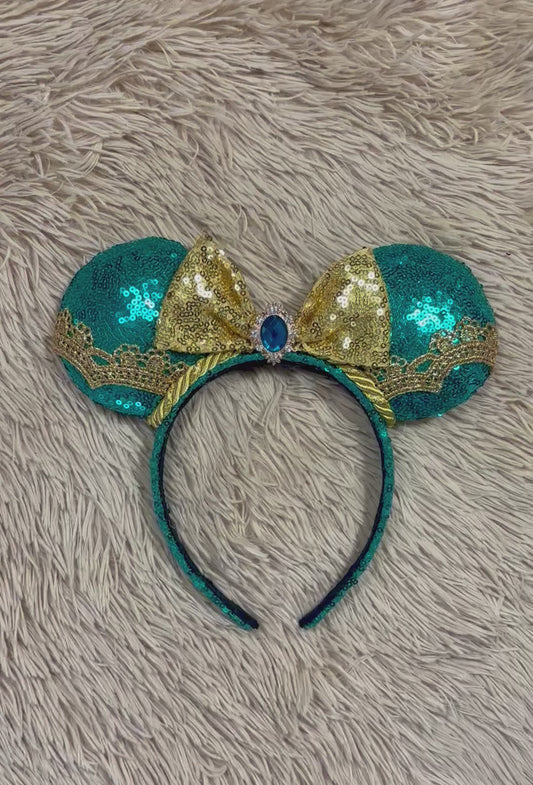 A pair of sequined mouse ears headband featuring teal and gold sequins, a large gold sequin bow with a blue gemstone center, and gold lace trim.