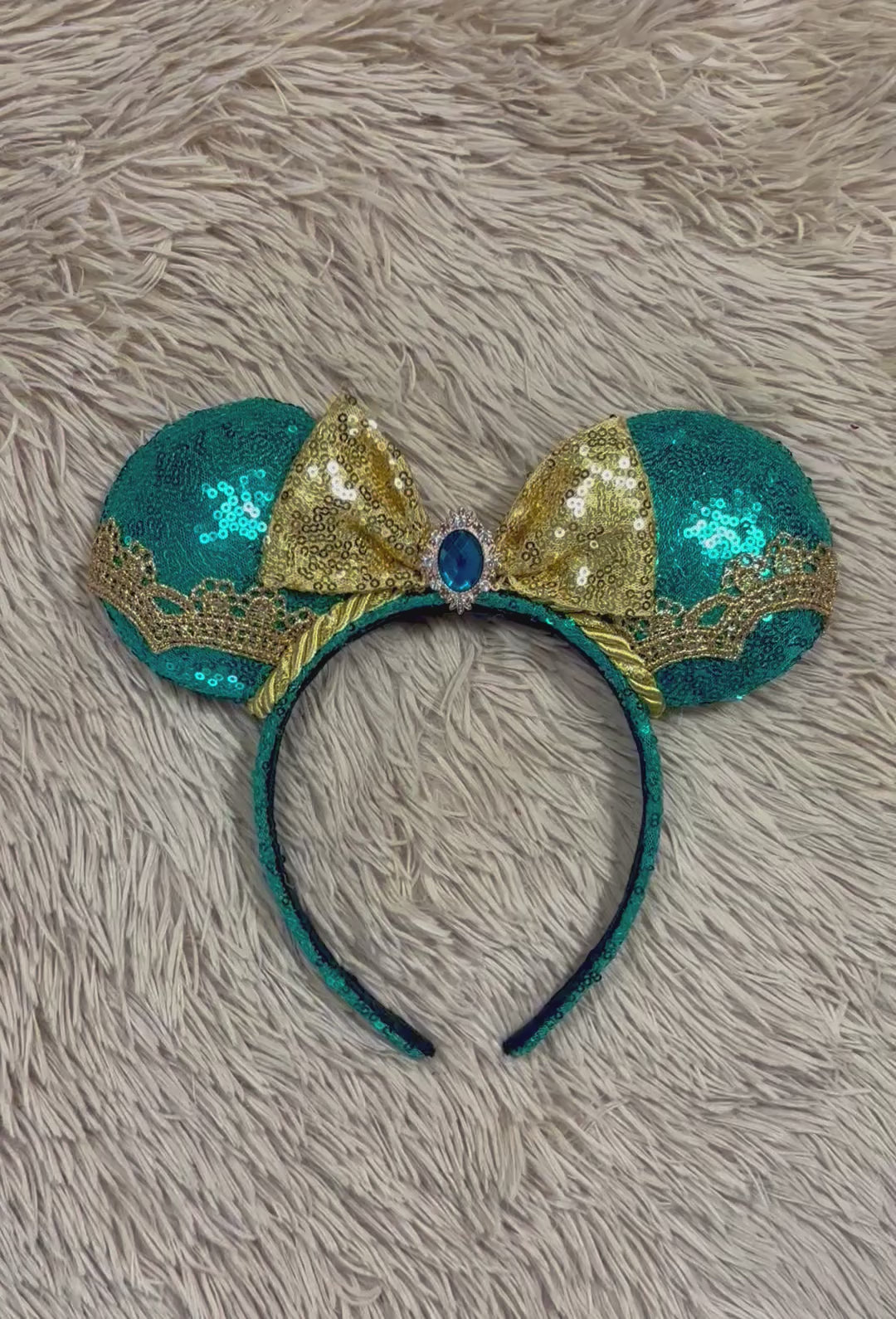 A pair of sequined mouse ears headband featuring teal and gold sequins, a large gold sequin bow with a blue gemstone center, and gold lace trim.