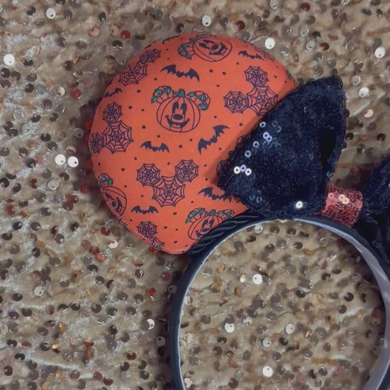 Halloween mouse ears with the fabric on both the front and back. It's orange with bats, mouse shaped pumpkins and spider webs. The headband is black universal sized and fits most. The set has a black with orange middle sequin bow. Item is shown on a gold sequin background.