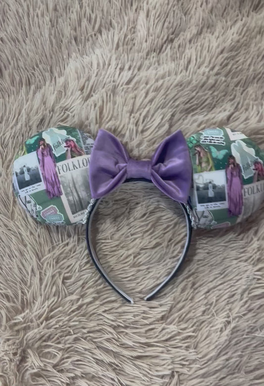 A pair of mouse-ear style headbands with a large purple bow in the center, The ears are covered in a collage print featuring vintage book pages, text, and illustrations of TS in a purple dress.