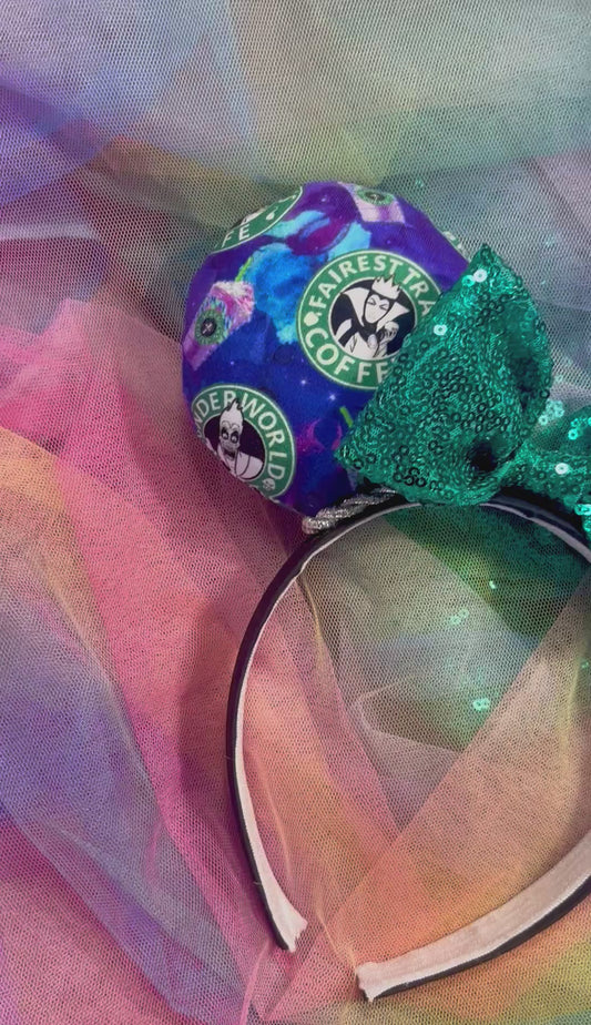 Colorful mouse ears with Coffee logo and villains on the fabric front and back with a teal sequin bow. Shown on a rainbow background.