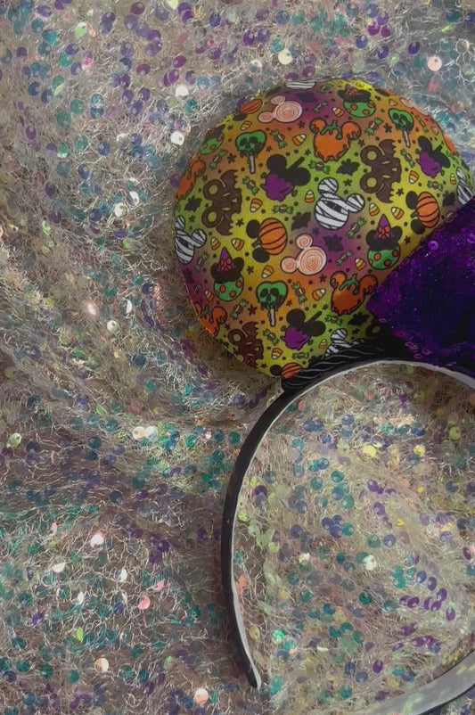 Colorful Halloween-themed mouse ears with a purple bow on a white glitter background