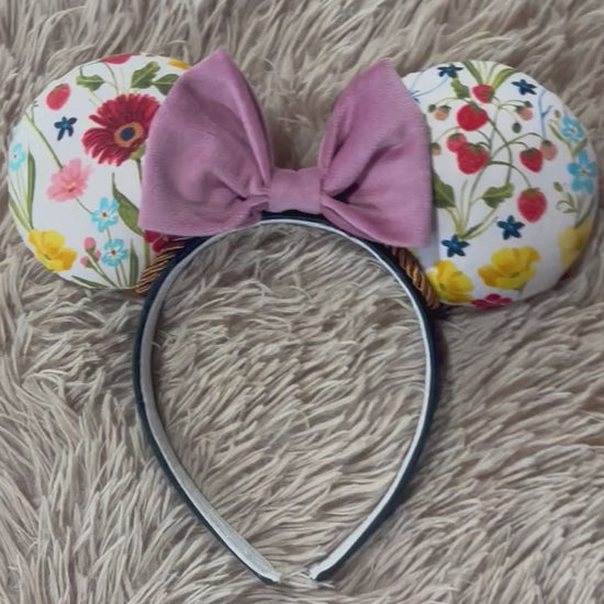 An ear headband accessory featuring a floral-themed print on the ears and a pink velvet bow in the center with copper colored twisted trim along the inner ears.
