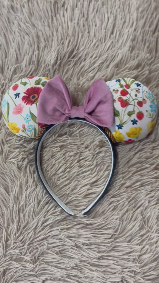 An ear headband accessory featuring a floral-themed print on the ears and a pink velvet bow in the center with copper colored twisted trim along the inner ears.
