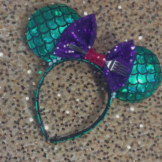Mermaid-themed headband with teal scales and a purple sequin bow with a red middle and a dinglehopper middle piece. Pearl garland trim along inner ears m on a matching mermaid scales headband.