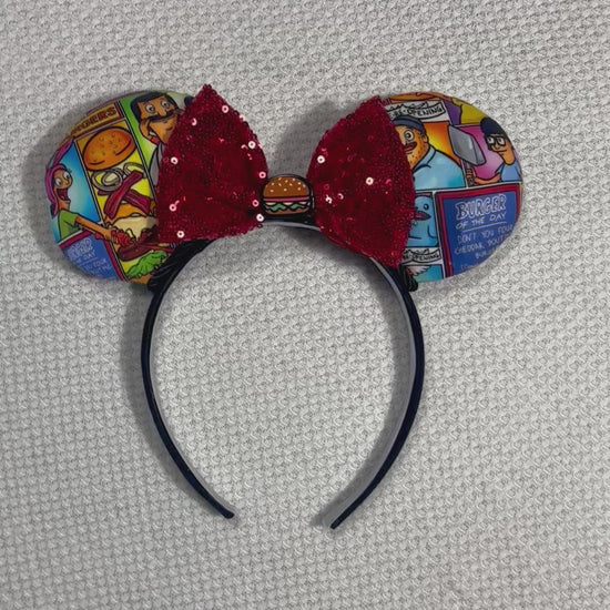 Handmade and ready to ship Belcher and friends mouse ears with the fabric on both the front and back. The headband is black universal sized and fits most. The set has a red sequin bow with a burger embellishment on the middle. Item is shown on a white background.