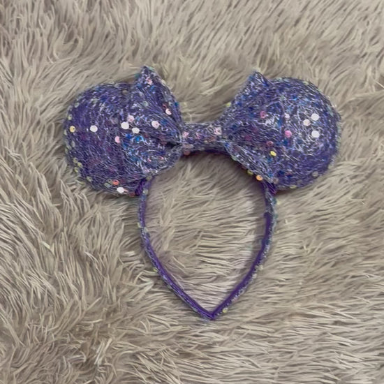 A purple sequined headband with large round ears and a bow, laid flat on a beige faux fur surface.