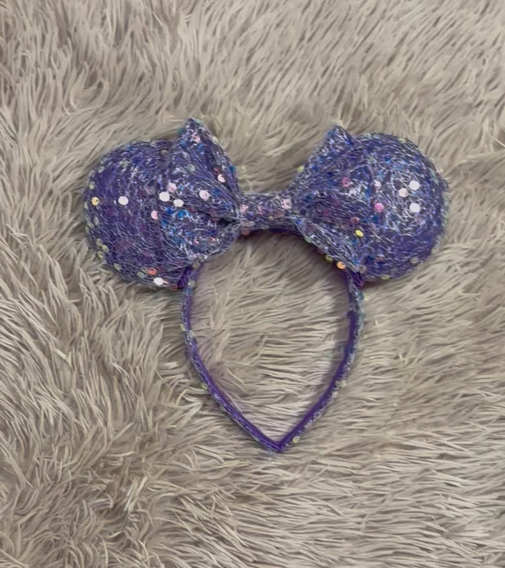 A purple sequined headband with large round ears and a bow, laid flat on a beige faux fur surface.